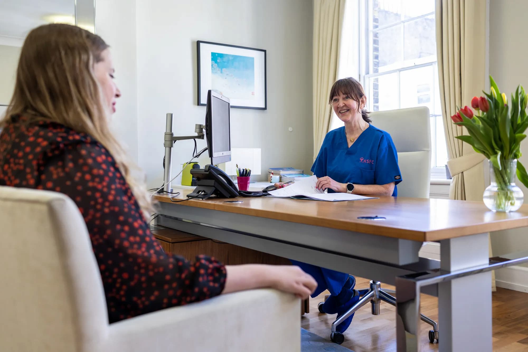 Dr Verstraete, consultant gynecologist, smiling and wearing scrubs, sitting at a desk and talking to a patient.