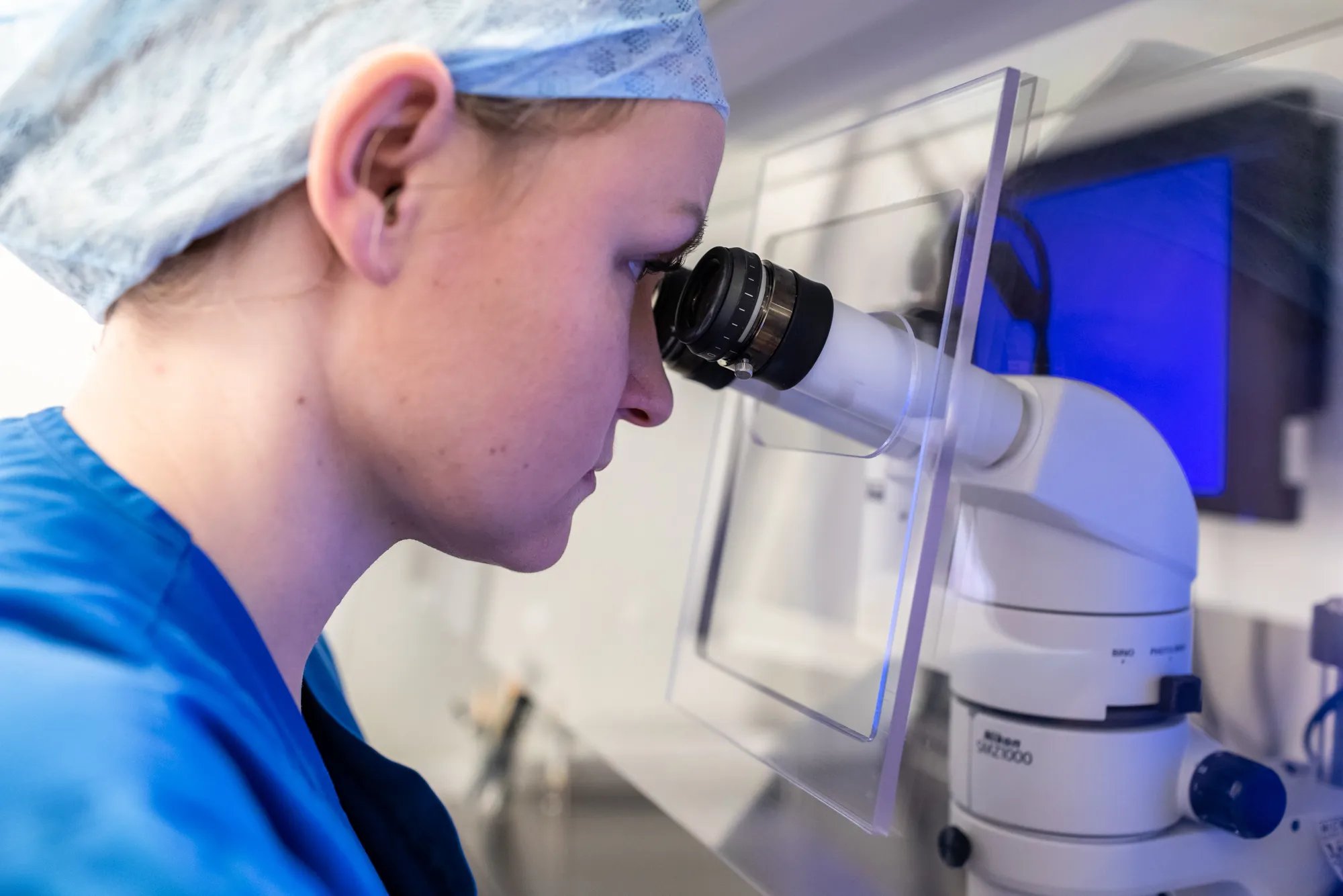 A close of an embryologist wearing scrubs and a hat, looking through a microscope. 